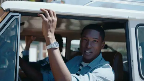 A man in a blue shirt rests his raised forearm near a yellow SUV window, showing a silver wristwatch beneath the white roofline.