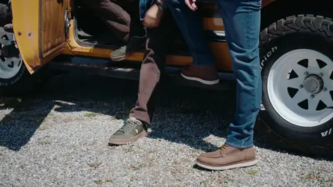 Brown leather shoes, green casual shoes, blue jeans and brown trousers are framed beside a yellow SUV running board and white wheel.