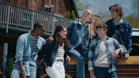Five people wearing coordinated denim and plaid gather beside a yellow vintage SUV outside a timber building.