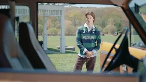 A man in a green striped rugby shirt stands on a field, seen through the open windows and brown seats of a vintage SUV.