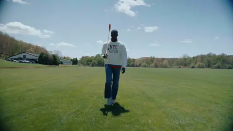 A man in a white graphic sweatshirt and blue jeans carries a polo mallet on a green field beneath a broad blue sky.