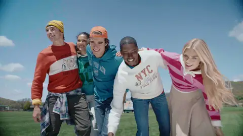 Five people in red, green, blue, white and pink layers lean together toward the camera beneath a clear blue sky.