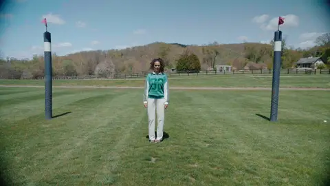 A woman in a green top and white trousers stands centered between tall striped polo goal posts on a grassy field.