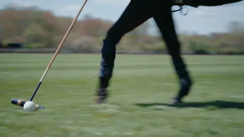 A wooden polo mallet meets a white ball on short grass beside the lower legs of a horse.