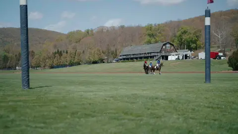 Mounted polo players cross a broad green field between striped goal posts, with wooded hills and a timber building beyond.