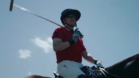 A polo rider in a red shirt, dark helmet and white trousers holds a mallet while seated on a horse against blue sky.