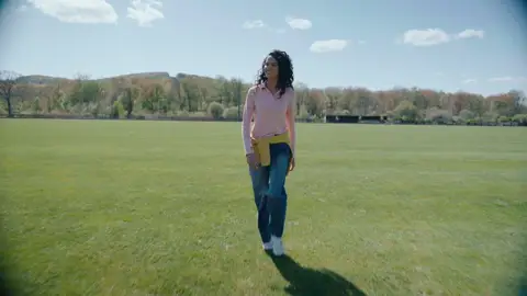 A woman in a pink top and wide blue jeans walks across grass with a yellow sweater tied at her waist.