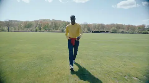 A man in a yellow shirt and dark blue jeans walks on a field with a red sweater tied around his waist.