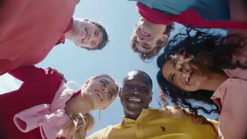 Five people in brightly colored polo shirts lean into a circle above a low camera, their faces framing the blue sky.