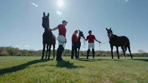 Three people in red tops stand with horses on a sunlit field, casting long shadows toward the low camera.