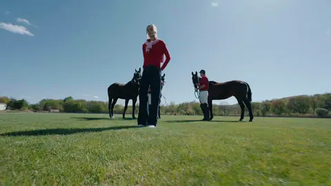 A woman in a red sweater, pink shoulder layer and wide blue jeans stands in the foreground while horses and handlers remain behind her.