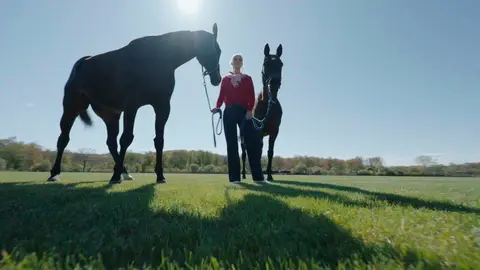 A woman in a red top holds the lead ropes of two brown horses on grass, with strong backlight and long foreground shadows.