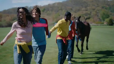 People in pink, blue, yellow and red layers form a hand-holding line beside a brown horse on a grassy field.