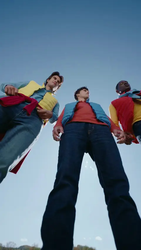 Three men in yellow, blue and red puffer vests and jeans stand above a low camera against blue sky in a vertical frame.