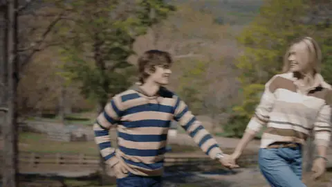 A man and woman in striped tops turn together while holding hands on a grassy hillside.