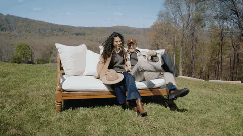 People in casual layers join a seated woman on a white-cushioned sofa outdoors with hills behind them.