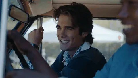 A passenger with a striped collar smiles and laughs behind the driver inside a vintage SUV.