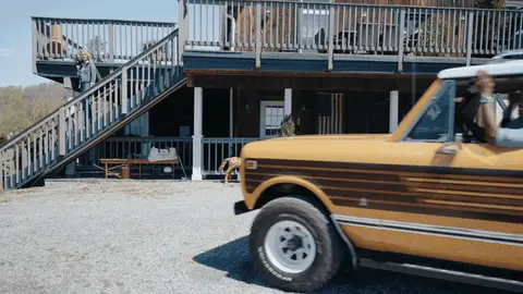 A yellow vintage SUV rolls into view beside a dark timber building on an outdoor drive.