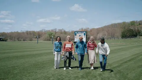 Five people in colorful casual layers walk together toward the camera across a green field.