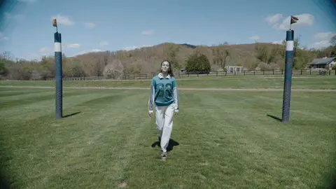 A woman in a green top and white trousers walks forward between striped polo goal posts on grass.