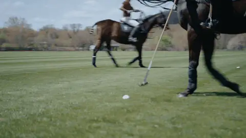 A polo mallet swings toward a white ball on the grass while a horse steps through the low-angle frame.