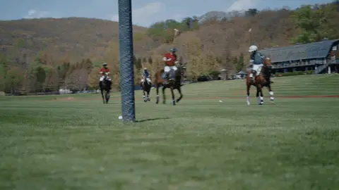 Polo riders in red and blue shirts approach on brown horses as the camera follows from a low angle.