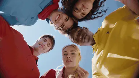Five people in colorful polo shirts lean together and smile around a low camera with blue sky in the center.