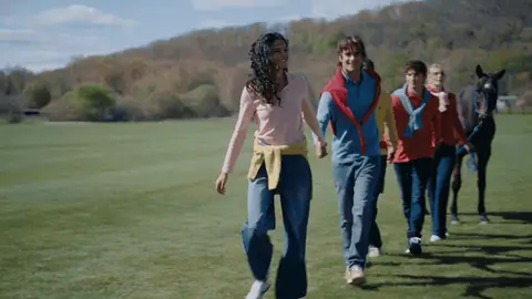 A colorful group steps forward and opens into a hand-holding line beside a brown horse on a grassy field.