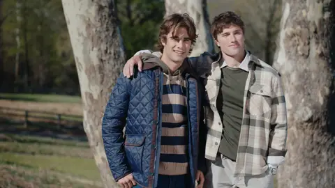 Two men stand with an arm around each other beside birch trees, wearing a navy quilted jacket and a cream-and-gray plaid overshirt.