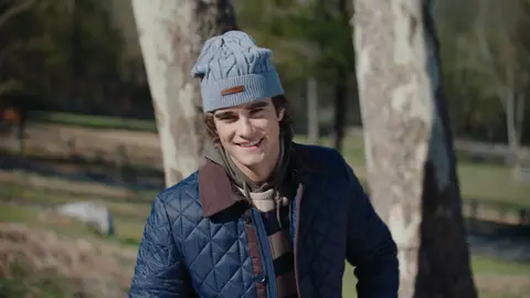 A smiling man wears a pale blue cable-knit beanie and navy quilted jacket beneath sunlit birch trees.
