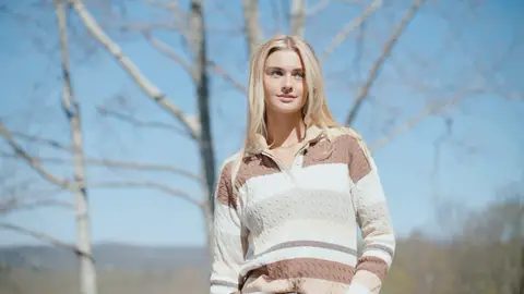 A blonde woman in a cream-and-brown striped knit sweater looks to the side with a slight smile, with bare trees and blue sky behind her.