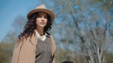 A woman with curly dark hair wears a tan felt hat, camel coat and dark cable-knit sweater vest against a sunlit hillside and blue sky.