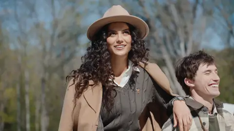 A woman in a tan hat and camel coat rests her arm on a smiling man in a gray plaid overshirt while friends sit beside them outdoors.