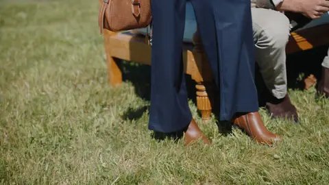 Brown ankle boots and navy wide-leg trousers stand beside a wooden outdoor sofa on grass, with another seated person's gray trousers and boots nearby.