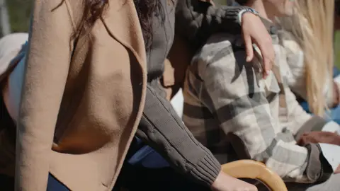 A close view shows a camel coat, dark ribbed knit sleeve and gray plaid overshirt, with a woman's arm resting across a seated man's shoulder.