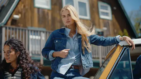 A blonde woman in a denim jacket and jeans sits beside the open window of a yellow vintage SUV, with another woman and a timber building nearby.