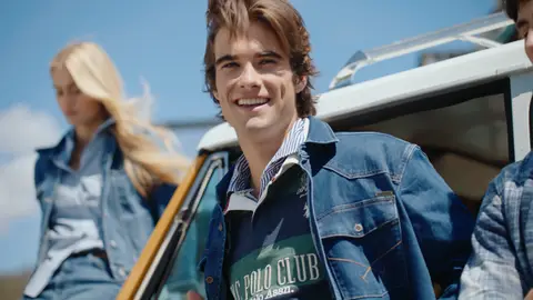 A smiling man wears a blue denim jacket over a green striped rugby shirt beside a yellow vintage SUV, with a seated woman softly blurred behind him.