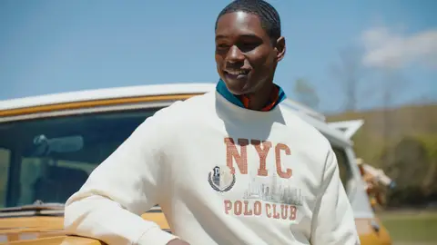 A smiling man in a cream NYC Polo Club sweatshirt rests his arm on a yellow vintage SUV in bright sunshine.