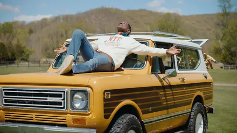 A man in a cream sweatshirt and jeans reclines with open arms on the hood of a yellow vintage SUV parked on a grassy field.