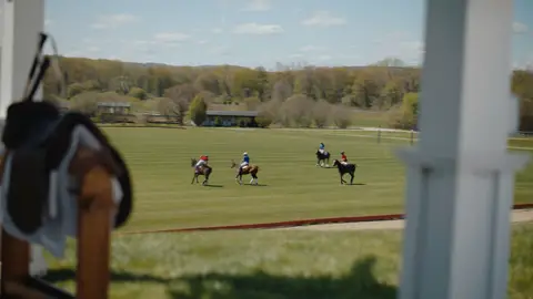 Mounted polo players cross a green field between white pavilion posts, with a saddle softly blurred in the foreground.