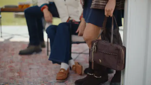 A brown handbag with whipstitched trim is held beside dark brown boots and a navy skirt in a white polo pavilion.