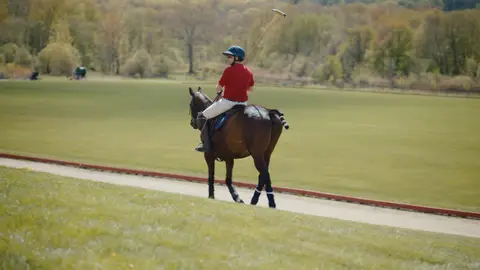 A helmeted polo rider in a red shirt and white trousers rides a brown horse along the edge of a green field beneath a wooded hillside.