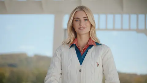 A smiling blonde woman wears a white cable-knit cardigan over blue and red collars beneath a white wooden polo pavilion.