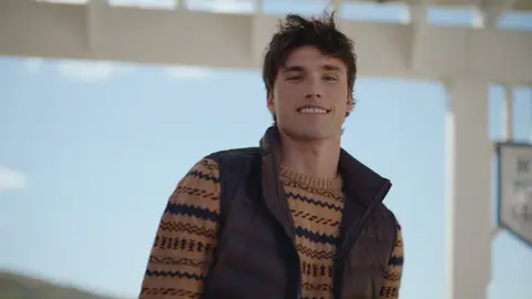 A smiling man wears a brown quilted vest over a tan-and-navy patterned sweater beneath a white polo pavilion.