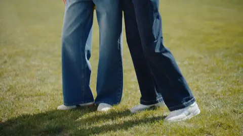 Two women stand side by side in wide-leg blue jeans and white sneakers on sunlit grass.