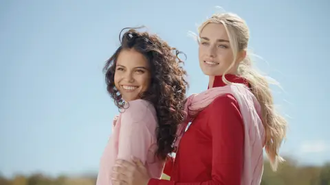 Two smiling women stand close together on a sunny field, wearing red and pale pink polo shirts with pastel sweaters draped over their shoulders and waist.