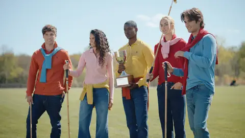 Five adults in orange, pink, yellow, red and blue polo shirts stand on a green field with a silver trophy and wooden polo mallets.