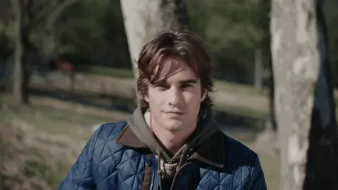 A man in a navy quilted jacket pulls a pale blue cable-knit beanie onto his head beside birch trees, then looks toward the camera.