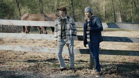 Two men wearing a navy quilted jacket and gray plaid overshirt walk beside a wooden paddock fence while a brown horse grazes behind them.
