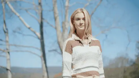A woman in a cream-and-brown striped sweater opens her arms and turns on a sunny hillside as her long blonde hair moves in the breeze.